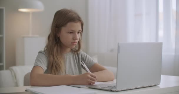 Menina adolescente está aprendendo distantemente, escrevendo em caderno de exercícios e olhando para a exibição de caderno, ficar em casa — Vídeo de Stock