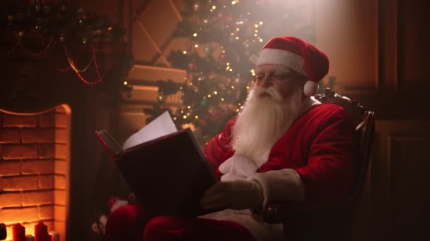 Joyful santa clausule zitten in zijn rocker in versierde kamer, het lezen van een boek met rode omslag - vakantiestemming, kerst geest concept. — Stockvideo