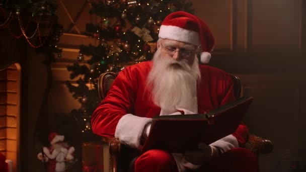 Γενειοφόρος ανώτερος άνθρωπος σε στολή ρήτρα santa flipping μέσα από σελίδες του βιβλίου με κόκκινο εξώφυλλο σε θεματικά διακοσμημένο δωμάτιο - Χριστούγεννα έννοια πνεύμα κοντά. — Αρχείο Βίντεο