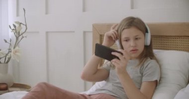 Liseli kız yatakta uzanıyor ve internet üzerinden akıllı telefon oyunu oynuyor, kulaklıkla müzik dinliyor.