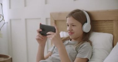 Neşeli küçük kız elinde akıllı telefonuyla yatakta uzanıyor ve oyun oynuyor, internette vakit geçiriyor.