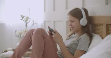 Genç kız akıllı telefonuyla iletişim kurmak için uygulama kullanıyor, kulaklıkla konuşuyor ve dinliyor, yatak odasında dinleniyor.