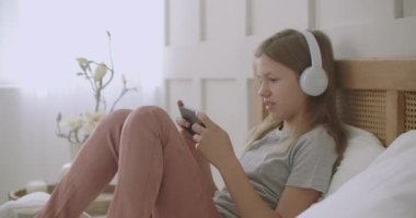 Liseli kız evde kalıyor ve telefonda oyun oynuyor, yatağa uzanıyor, kulaklıkla müzik dinliyor.