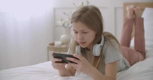 Menina adolescente está deitada na cama, falando e usando o aplicativo no smartphone, conversando on-line com amigos, entretenimento e lazer de crianças em férias e fim de semana — Vídeo de Stock