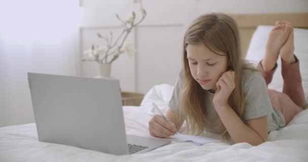 Niña está dibujando y escribiendo en copybook, acostado en la cama delante de la computadora portátil en su habitación, esperando lección en línea — Vídeos de Stock