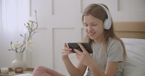 Klein meisje is met behulp van smartphone en draadloze hoofdtelefoon zitten op bed in de kamer, het spelen van spel en glimlachen — Stockvideo