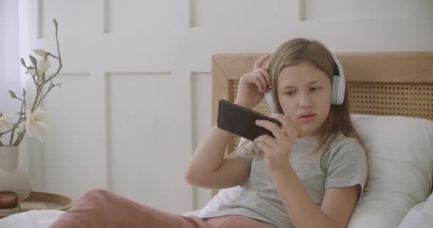 Schoolmeisje ligt op bed in de kamer en het spelen van spel op de smartphone via internet, het luisteren naar muziek met een koptelefoon — Stockvideo