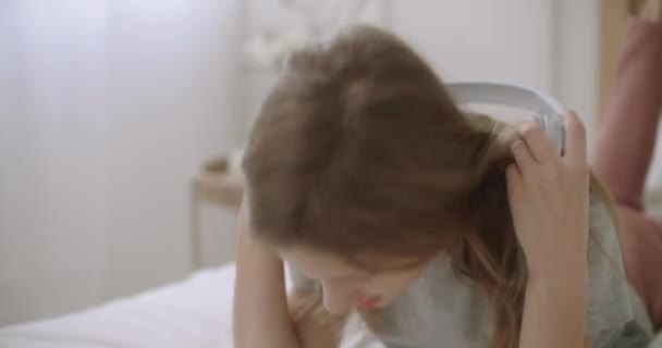Menina está colocando fones de ouvido sem fio e levando smartphone para ouvir música e usar a internet, deitado na cama — Vídeo de Stock