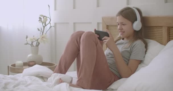 Niña está utilizando el teléfono inteligente, jugando juego en línea después de despertarse en la mañana del domingo, adicción a Internet de los escolares — Vídeo de stock