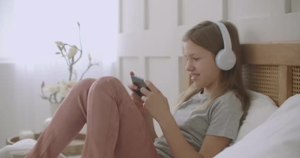 Mooi meisje is met behulp van app voor communicatie op de smartphone, praten en luisteren met een koptelefoon, rusten in haar slaapkamer — Stockvideo