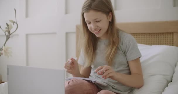 Menina adolescente está se preparando para a aula on-line por internet, segurando copybook, sentado na cama na frente do laptop — Vídeo de Stock