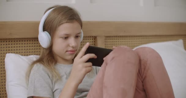 Écolière regarde drôle de vidéo sur l'affichage de smartphone et écouter par écouteurs dans sa chambre, regarder des dessins animés et des films — Video