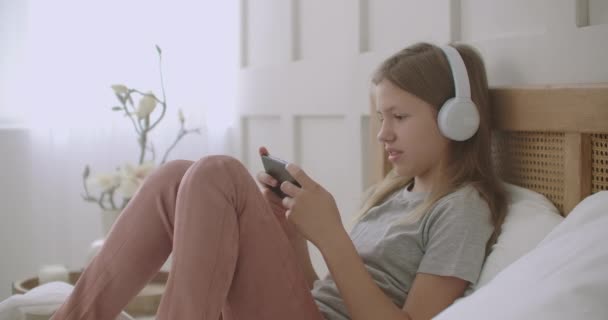 Schoolmeisje blijft thuis en spelen spel in de telefoon, liggend op bed, luisteren naar muziek met een koptelefoon — Stockvideo