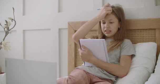 子供のためのオンライン教育、女の子は自宅から学び、ノートパソコンでベッドに座り、コピーブックとペンを持ってレッスンを受けます — ストック動画