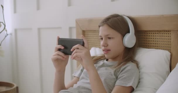 Schoolmeisje is het spelen van video game in smartphone via internet, met behulp van een koptelefoon en liggend op bed tijdens de vakantie — Stockvideo