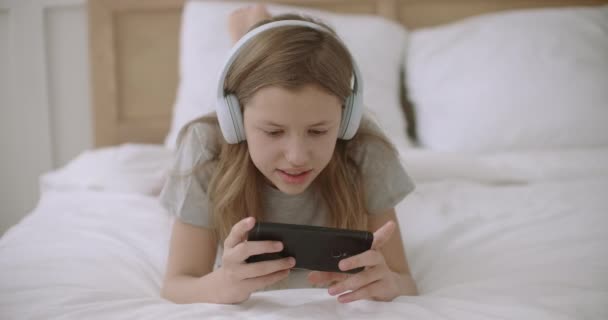 한 십 대 소녀가 스마트폰으로 인터넷 서핑을 하며 사이트의 정보를 읽고 방과 후 집에서 침대에 누워 있습니다. — 비디오
