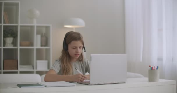 Menina está aprendendo on-line, se comunicando com o professor pela internet, usando fones de ouvido e laptop, fazendo lição de casa no caderno de exercícios — Vídeo de Stock