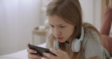 Liseli kız akıllı telefonuyla internette sörf yapıyor ve ekrandaki resimlere bakıyor, evdeki yatakta uzanıyor, sensör ekranını karıştırıyor ve tıklatıyor.
