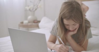 Liseli kız dizüstü bilgisayarı ve defteriyle yatakta uzanıyor, ödev yazıyor ve dizüstü bilgisayarına bakıyor.