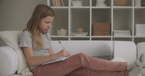 Tienermeisje is schrijven in copybook, zitten thuis in de woonkamer op de bank in het weekend, ontspannen tijdens vakantie of weekend — Stockvideo