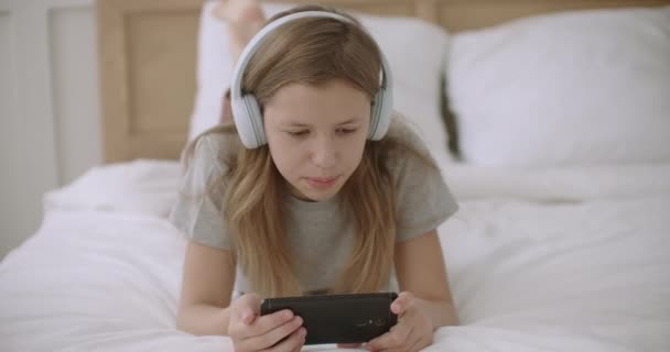 Gioiosa teen girl sta leggendo il testo sullo schermo del telefono cellulare, sdraiato sul letto, utilizzando cuffie wireless e wifi — Video Stock