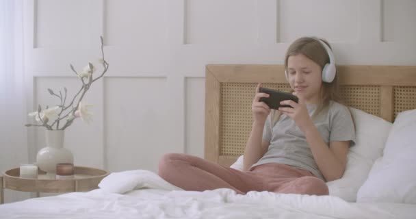 Kind meisje is het spelen van spel in smartphone na schoollessen, zitten op bed alleen thuis, jeugd — Stockvideo