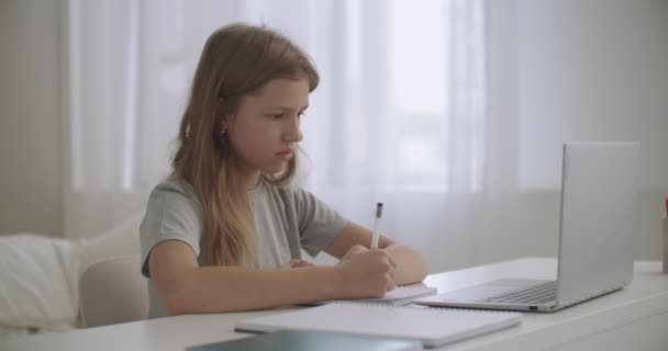 Menina adolescente está aprendendo distantemente em casa, olhando para a tela do laptop e escrevendo em caderno de exercícios — Vídeo de Stock