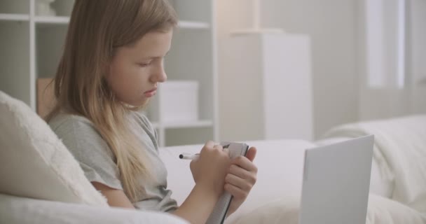 Nastolatka dziewczyna słucha wykładu online nauczyciela przez laptop w domu, pisanie w copybook, e-learning dla uczniów podczas epidemii — Wideo stockowe