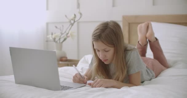 Colegiala está charlando en línea por ordenador portátil desde casa, acostado en la cama, hablando alegremente y dibujo en copybook — Vídeos de Stock
