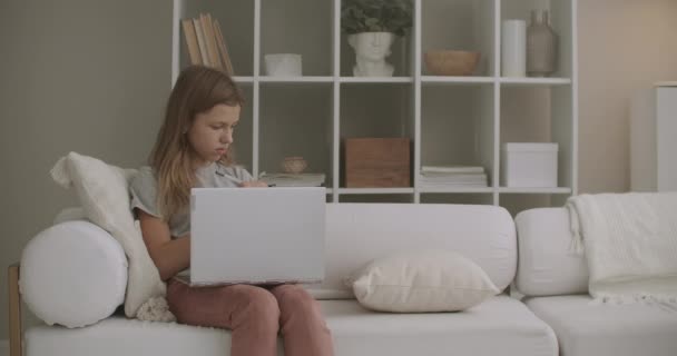 Gadis kecil bekerja dengan notebook dan menggambar di buku fotokopi di rumah, duduk di sofa di ruang tamu pada akhir pekan — Stok Video