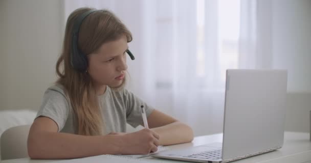 Niña está aprendiendo en línea por Internet, escuchar al profesor a través de los auriculares y mirar la pantalla del cuaderno, escribir notas en el libro de ejercicios — Vídeos de Stock