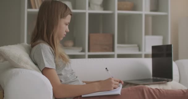 Genç kız okuldan sonra ev ödevi yapıyor, egzersiz kitabına yazıyor, kanepede oturuyor, okul çocukları için e-öğrenim yapıyor. — Stok video