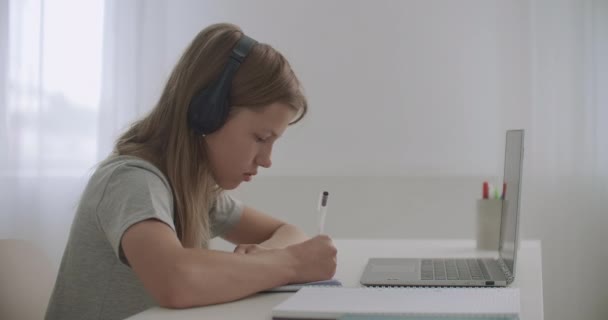 Schülerin macht Hausaufgaben, sitzt am Tisch in ihrem Zimmer, benutzt Laptop und Kopfhörer, schreibt in Schulheft — Stockvideo