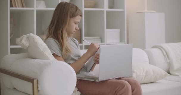 Genç kız evde dizüstü bilgisayarla eğitim görüyor ve uzaktan öğreniyor, defterine notlar yazıyor, rahatlıyor ve ev ortamında — Stok video