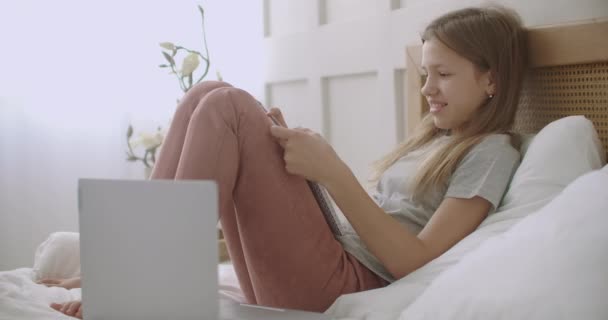 Glad skolflicka pratar och ler, sitter på sängen med copybook efter online-lärande session, e-lärande — Stockvideo