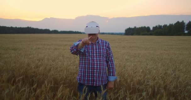 Uma fazenda de idosos usa um capacete VR enquanto está em pé em um campo ao pôr do sol. Usar gestos para a realidade virtual na agricultura. — Vídeo de Stock
