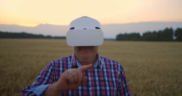 Una granja de ancianos utiliza un casco VR mientras está de pie en un campo al atardecer. Utilizar gestos para la realidad virtual en la agricultura. — Vídeos de Stock