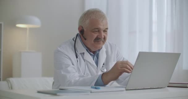 Amable médico alegre está consultando en línea, trabajando por Internet con el paciente, charlando por videollamada desde su computadora portátil, telemedicina moderna — Vídeo de stock