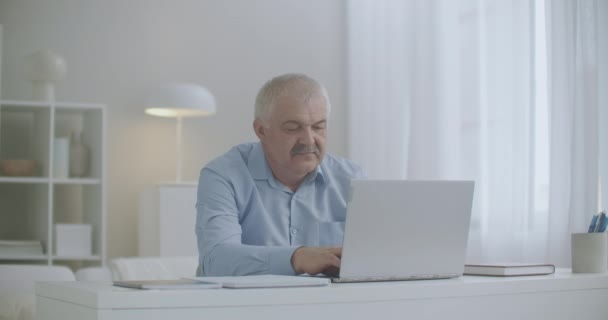 Baş ağrısı olan yorgun orta yaşlı bir adam evde dizüstü bilgisayarla çalışıyor ve aşırı uzatmadan dolayı sefalji, gerilim tipi baş ağrısı çekiyor. — Stok video