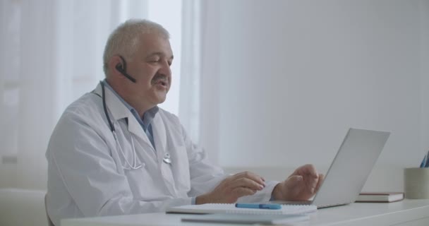 医師はオンラインでコンサルティングをしています私立病院のオフィスに座って患者と話すためにラップトップを使用しています遠隔医療技術 — ストック動画