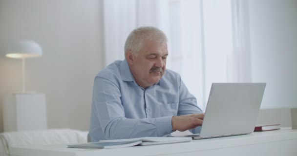 Aged gray haired man is working remotely from home, communicating by chat, typing message on notebook with internet connection — Stock Video