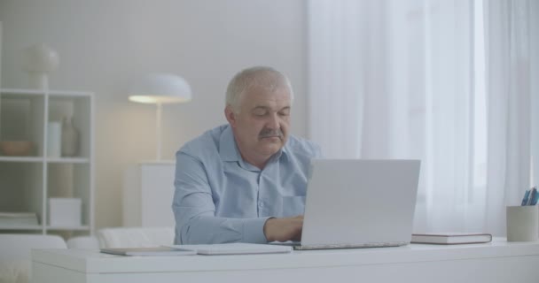 Aged man is stretching hands during working with laptop at home, feeling overextension and fatigue of muscles, feeling bad at workplace — Stock Video