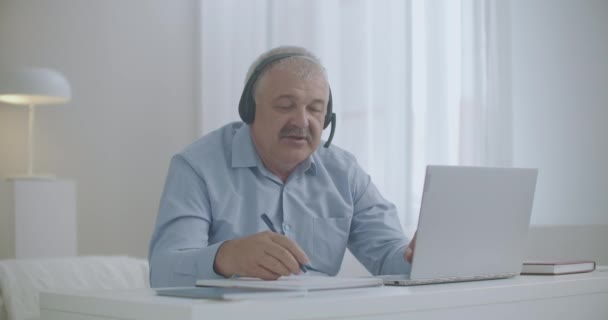 Aged man is working from home, using notebook for communicating with colleagues and partners, making notes on paper — Stock Video