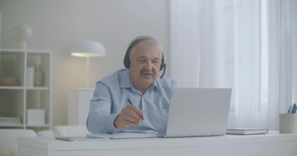 Middle-aged man is communicating by online chat with colleagues and employees from home using laptop and headphones — Stock Video