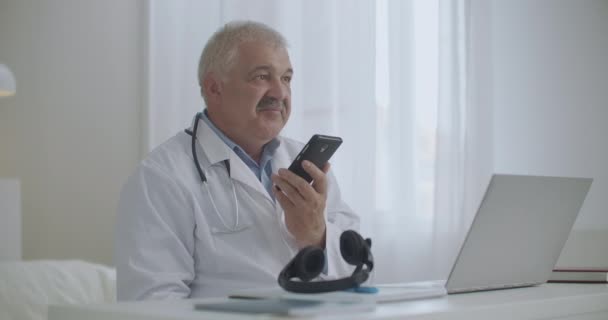 Врач-мужчина разговаривает по громкой связи в мобильном телефоне в своем кабинете в клинике, записывает голосовое сообщение, консультирует пациента по мобильному телефону — стоковое видео