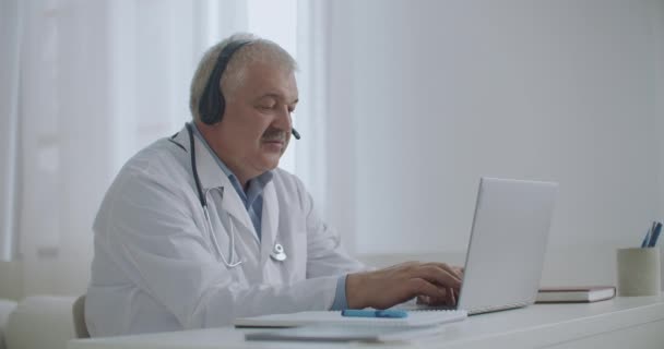 Man läkare talar med patienten på nätet genom videosamtal på laptop, lyssna med hörlurar och fylla medicinsk rapport arbetar på distans — Stockvideo