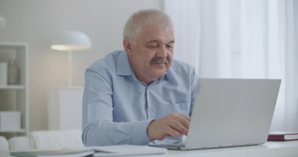 Middle-aged man with moustache is typing text on keyboard of laptop, browsing internet sites, working with notebook from home — Stock Video