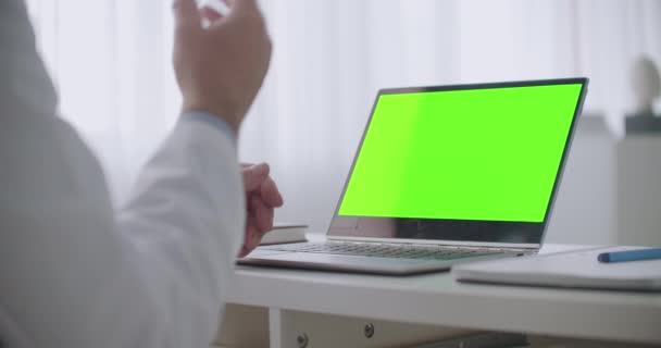 Online konzultace s lékařem, notebook se zelenou obrazovkou pro chroma klíčovou technologii na stole a gestikulující ruce lékaře — Stock video