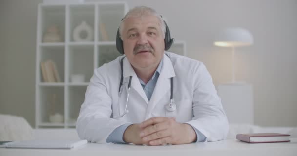 Męski lekarz rozmawia z kolegami lub pacjentami przez połączenie wideo, siedzenie w klinice, zdalna komunikacja podczas pandemii koronawirusa — Wideo stockowe