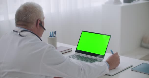 Αρσενικό γιατρός συμβουλεύεται σε απευθείας σύνδεση από το γραφείο του, προβολή σε φορητό υπολογιστή με πράσινη οθόνη για την τεχνολογία κλειδί chroma, κοιτάζοντας την οθόνη — Αρχείο Βίντεο